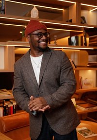 Blazer texturé marron sur une chemise blanche, associé à un jean foncé. La personne porte un bonnet rouge en tricot et des lunettes rondes, les mains jointes.