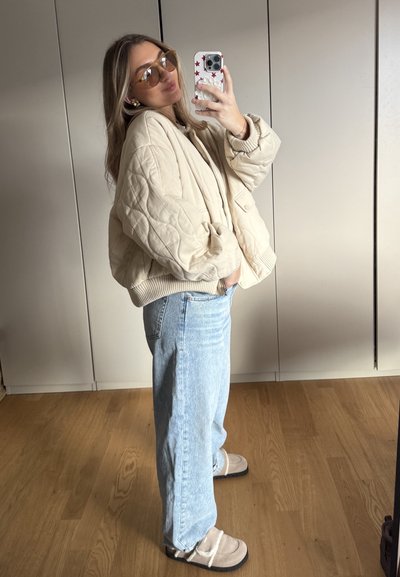Chaqueta bomber acolchada beige con puños de canalé, jeans de pierna ancha azul claro y zapatos beige tipo slip-on con correas, estilizados con gafas de sol de gran tamaño.