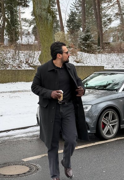 Hombre con abrigo negro y guantes sosteniendo una taza de café, caminando por una calle nevada junto a un coche gris y árboles en un entorno invernal.