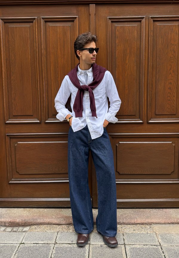 Homme portant des lunettes de soleil, une chemise blanche, un pull bordeaux noué sur les épaules, un jean large bleu et des chaussures marron, debout devant des portes en bois.