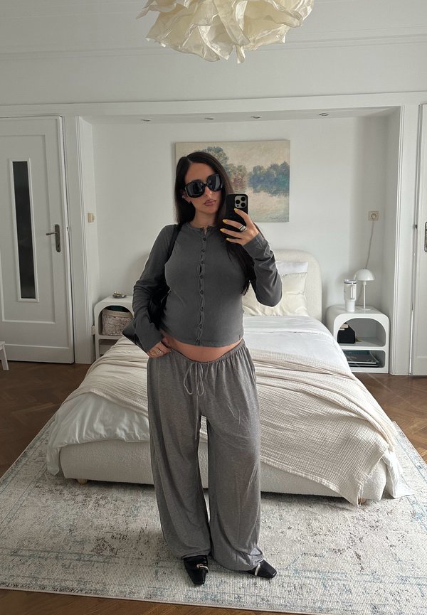 Nainen, jolla on tummat aurinkolasit ja harmaat kotivaatteet, ottaa selfien makuuhuoneessa, jossa valkoinen sänky, yöpöydät ja seinämaalaus.
