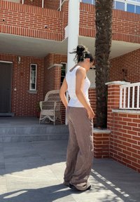 Top bianco senza maniche, pantaloni ampi a righe beige e marroni, calzature nere, in piedi su un patio piastrellato vicino a un muro di mattoni e a una palma.