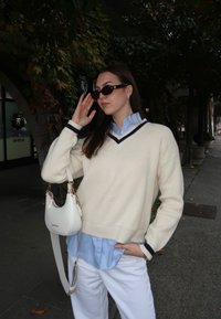 Béžový pletený sveter s námorníckymi modrými akcentmi, vrstvený cez svetlomodrú košeľu. Biela kabelka so zlatou kovovou výbavou, kombinovaná s bielymi nohavicami.