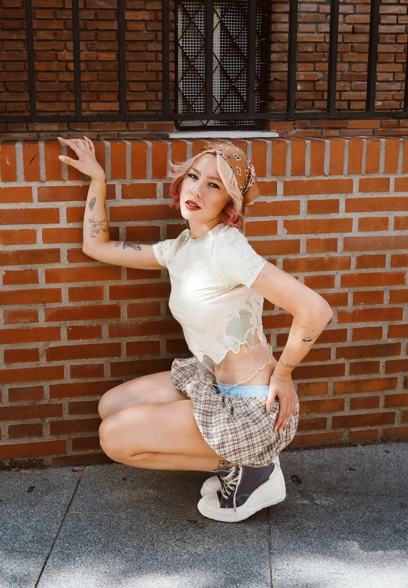Jeune femme tatouée portant un foulard, un haut blanc transparent, une jupe à carreaux et des baskets, accroupie contre un mur de briques en plein air.