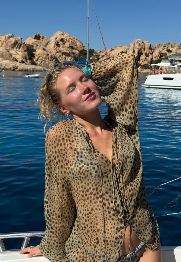 Frau in einer durchsichtigen Bluse mit Leopardenmuster lehnt sich an das Geländer eines Bootes, die Augen geschlossen, von der Sonne beleuchtet, mit felsigen Klippen und Wasser im Hintergrund.