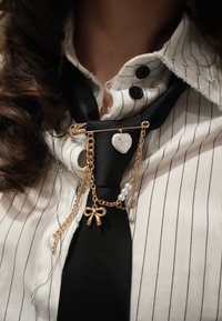 Gros plan sur une cravate noire avec une épingle de sûreté dorée décorative tenant un cœur, un nœud, des perles et des chaînes de strass sur une chemise blanche à rayures fines.