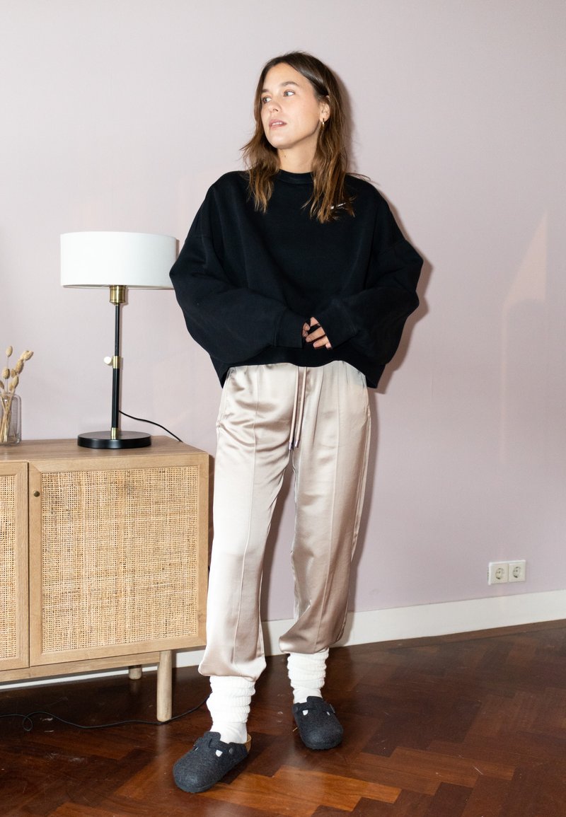 Mustat suurikokoiset collegepaidat, kermaväriset satiinipöksyt narulla, ja mustat tossut valkoisilla yksityiskohdilla. Huone, jossa on puulattia ja lamppu.