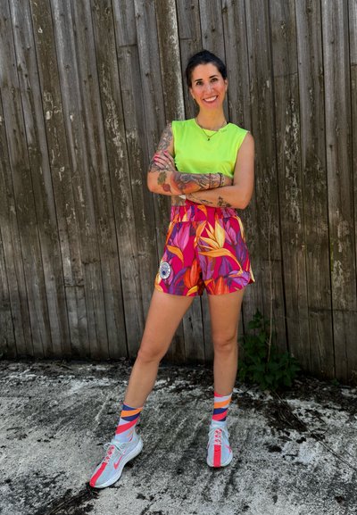 Mujer con tatuajes que lleva una camiseta verde neón, pantalones cortos florales coloridos, calcetines de estampado y zapatillas deportivas, de pie con los brazos cruzados junto a una cerca de madera.