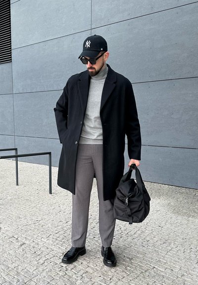 Hombre con gorra negra, gafas de sol, abrigo negro, jersey de cuello alto gris, pantalones grises, zapatos negros, sosteniendo una bolsa negra, de pie sobre un pavimento de piedra.
