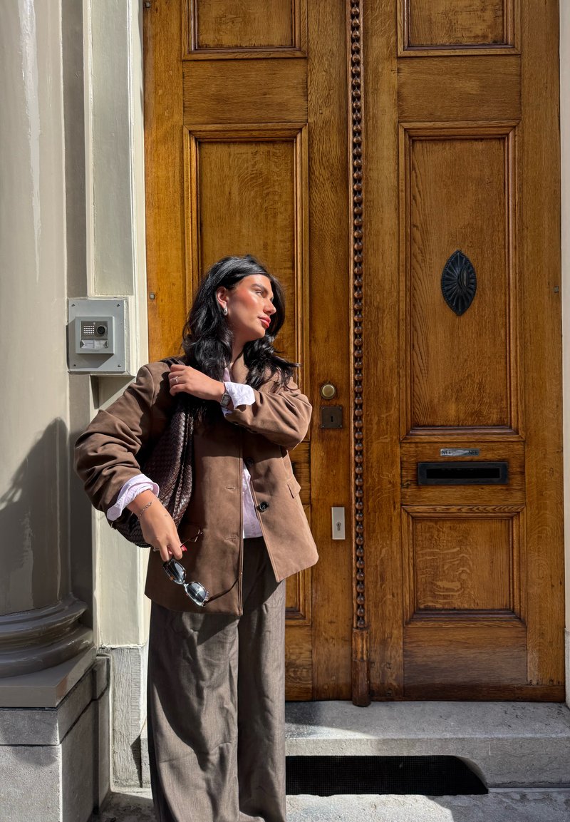 Braune Wildlederjacke über einem hellen Hemd, kombiniert mit lockeren braunen Hosen. Halte eine große geflochtene Tasche und Sonnenbrillen, stehe neben einer Holztür.