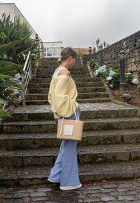 Sweater oversized amarelo claro, calças largas azuis, segurando uma bolsa de palha com um detalhe branco, a standing em degraus de pedra.