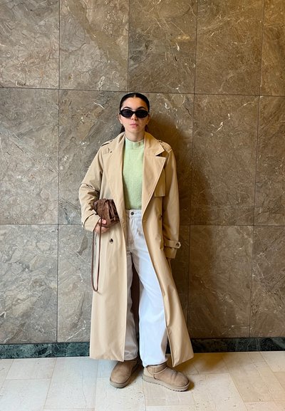 Gabardina beige, suéter verde claro, pantalones blancos, botas color tostado y bolso pequeño marrón; gafas de sol oscuras; de pie contra una pared de mármol.