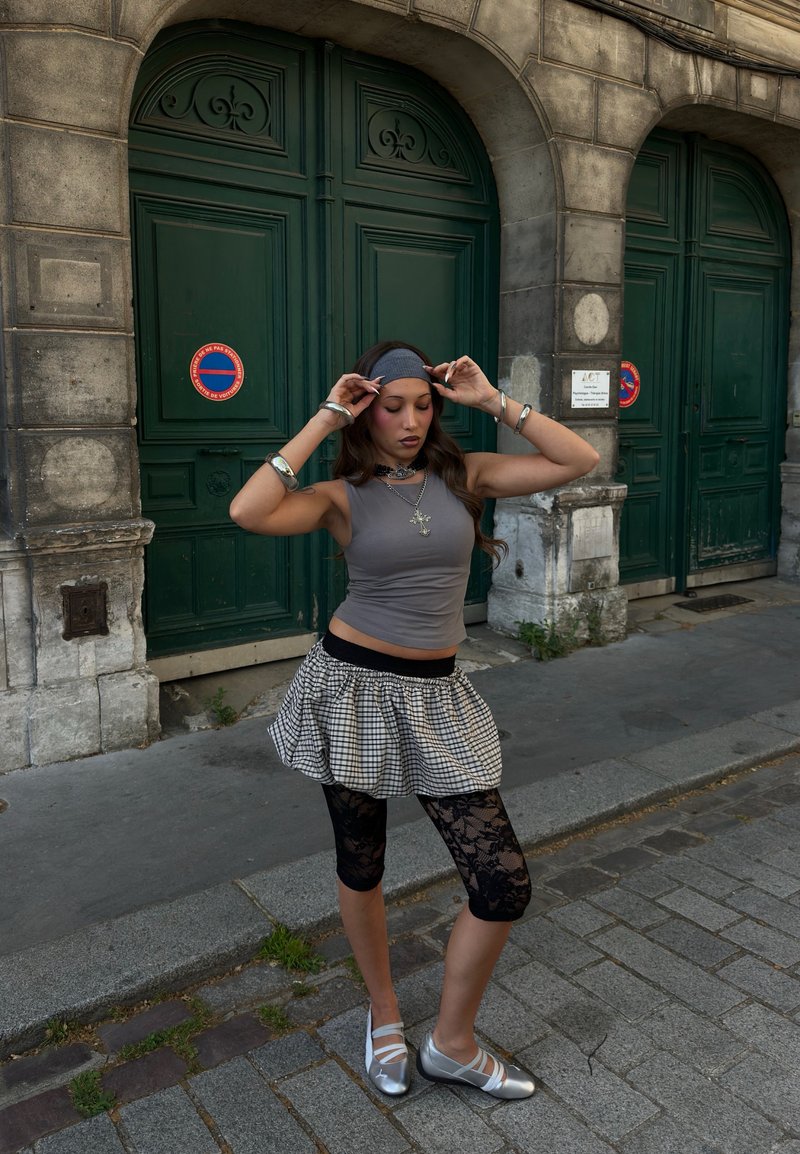 Femme en haut sans manches gris, jupe à carreaux, leggings en dentelle noire et chaussures argentées ajustant un serre-tête noir sur un trottoir en ville avec des portes vertes derrière.