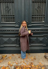 Ung kvinna med långt blont hår som bär en brun kappa, blå jeans och stövlar står vid en utsmyckad svart dörr på en höstpromenad med fallna löv.