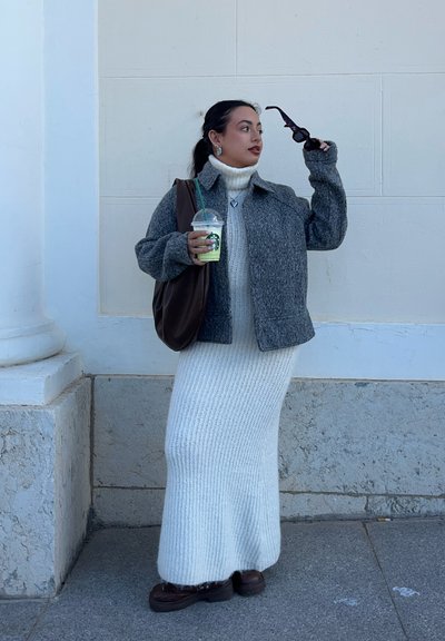 Mujer con chaqueta gris y vestido largo de punto blanco sosteniendo gafas de sol y una bebida de Starbucks, de pie contra una pared de piedra clara al aire libre.