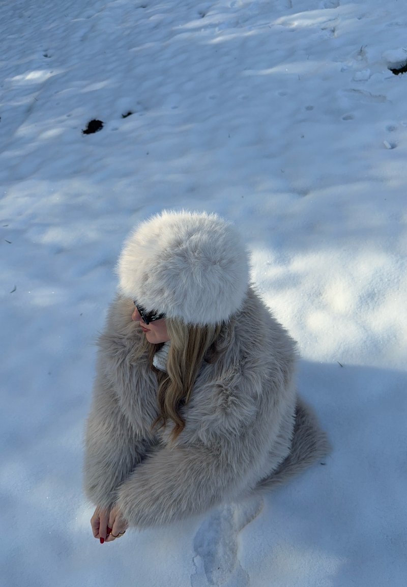 Manteau en fausse fourrure beige clair, coupe oversize avec texture longue et ébouriffée ; chapeau blanc moelleux assorti ; assis dans la neige avec des lunettes de soleil noires.