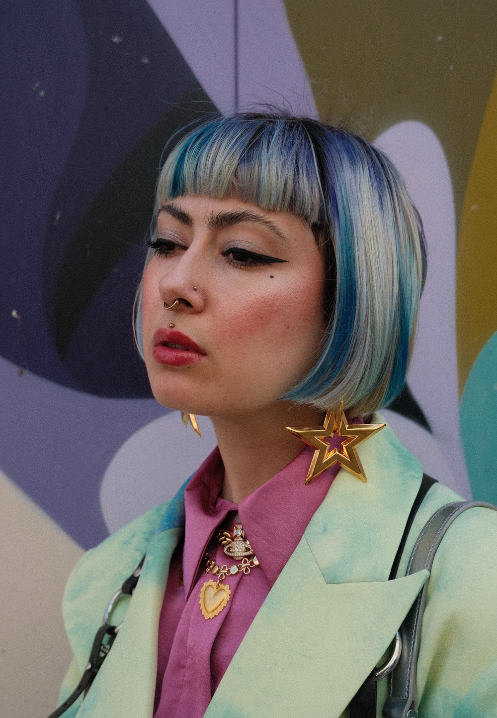 Ένα άτομο με μαλλιά σε μήκος ώμου σε μπλε και ξανθό χρώμα φοράει ένα μέντα πράσινο σακάκι πάνω από ένα ροζ πουκάμισο. Είναι ορατά εντυπωσιακά σκουλαρίκια και κοσμήματα σε σχήμα χρυσού αστεριού.