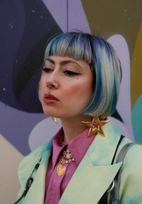 Ένα άτομο με μαλλιά σε μήκος ώμου σε μπλε και ξανθό χρώμα φοράει ένα μέντα πράσινο σακάκι πάνω από ένα ροζ πουκάμισο. Είναι ορατά εντυπωσιακά σκουλαρίκια και κοσμήματα σε σχήμα χρυσού αστεριού.