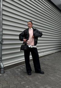 Μαύρο μπουφάν, ροζ πλεκτό πουλόβερ πάνω από λευκή πουκαμίσα, φαρδιά μαύρα παντελόνια και μικρή μαύρη τσάντα. Φόντο από υφασμάτινο γκρι τοίχο.