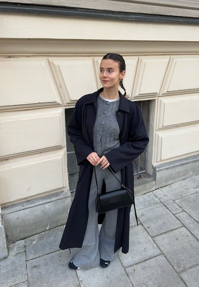 Largo abrigo azul marino sobre un cárdigan gris claro con botones, pantalones gris de pierna ancha y bolsa negra. Zapatos negros planos y accesorios mínimos.
