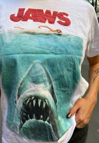 Weißes T-Shirt mit einem Grafikdesign eines Weißen Hais mit geöffnetem Maul, überlagert von einem Schwimmer, und dem Text "JAWS" in fettem Rot oben.