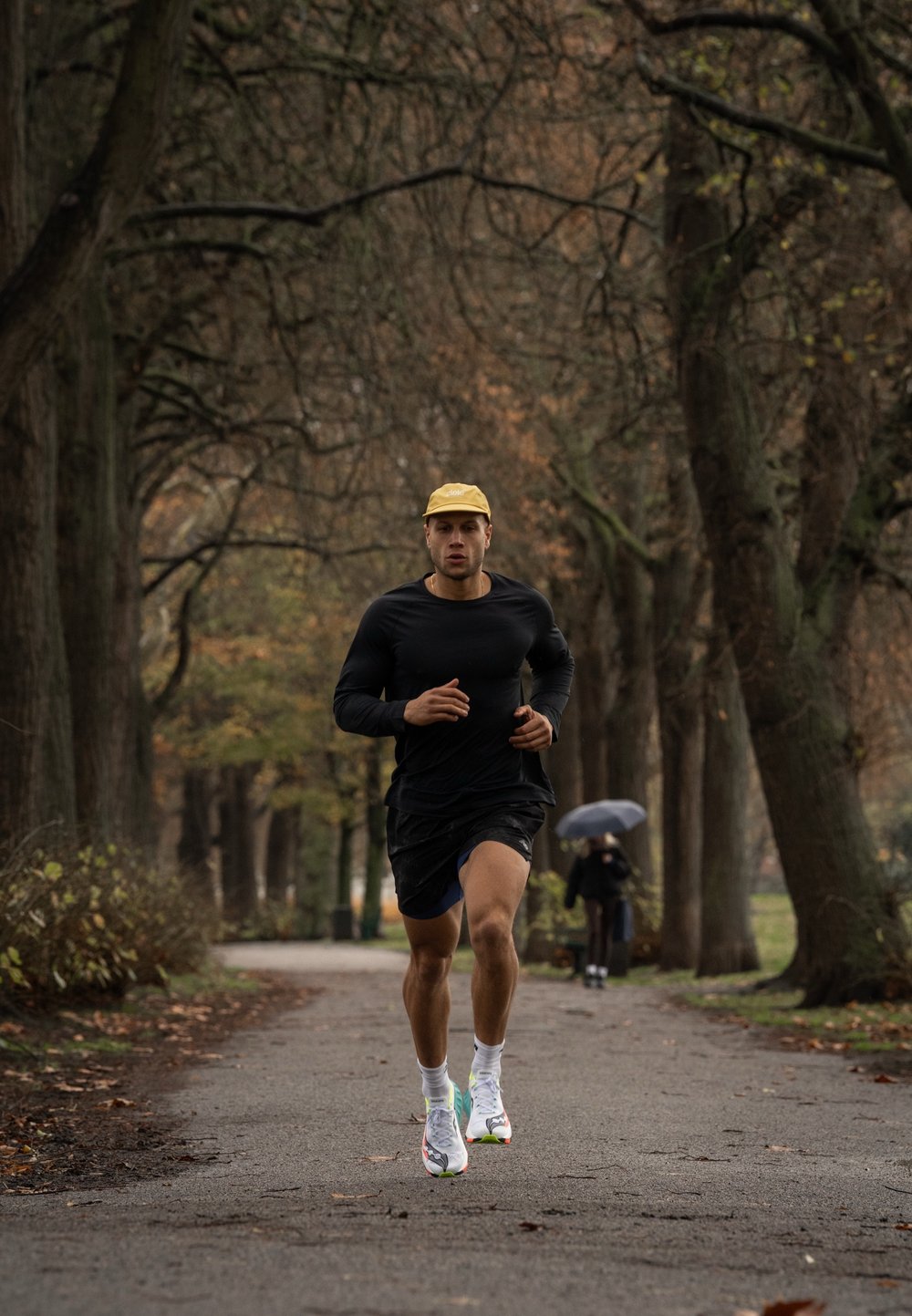 Atlét nosiaci čierne tričko s dlhým rukávom a čierne šortky, bežiaci po ceste lemovanej stromami. Biele a farebné športové topánky, žltá čiapka.