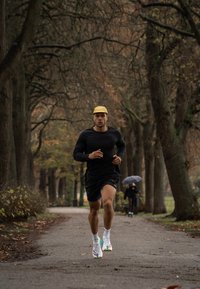 Atlét nosiaci čierne tričko s dlhým rukávom a čierne šortky, bežiaci po ceste lemovanej stromami. Biele a farebné športové topánky, žltá čiapka.