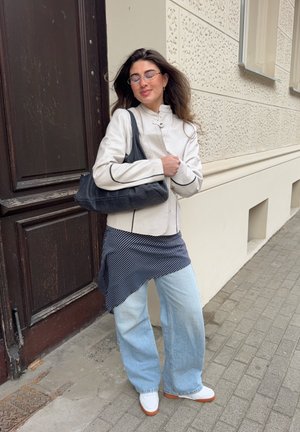 Mujer de cabello largo con gafas, chaqueta blanca, falda a rayas, vaqueros azul claro, zapatos blancos y bolso negro, de pie junto a una pared texturizada de un edificio.