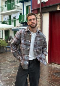 Vilnas pleda krekls pelēkā, brūnā un zilā toņos ar apkakli, pogām priekšpusē un garām piedurknēm, valkāts pār baltu T-kreklu un tumšām biksēm.