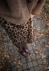 Person wearing brown fuzzy jacket, leopard print skirt, and pointed dark brown boots standing on leaf-strewn tiled pavement in autumn.