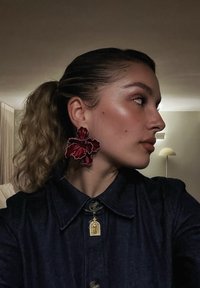 Frau mit lockigem Pferdeschwanz, die große rote Blumenohrringe, ein dunkles Hemd mit Kragen und eine goldene Anhänger-Halskette trägt, in einem sanft beleuchteten Raum.