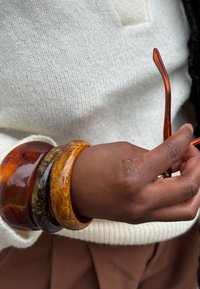 Una mano di pelle scura tiene occhiali da sole con tonalità ambra, indossando tre braccialetti di resina spessi nei toni marrone e oro sopra un maglione di maglia bianca.
