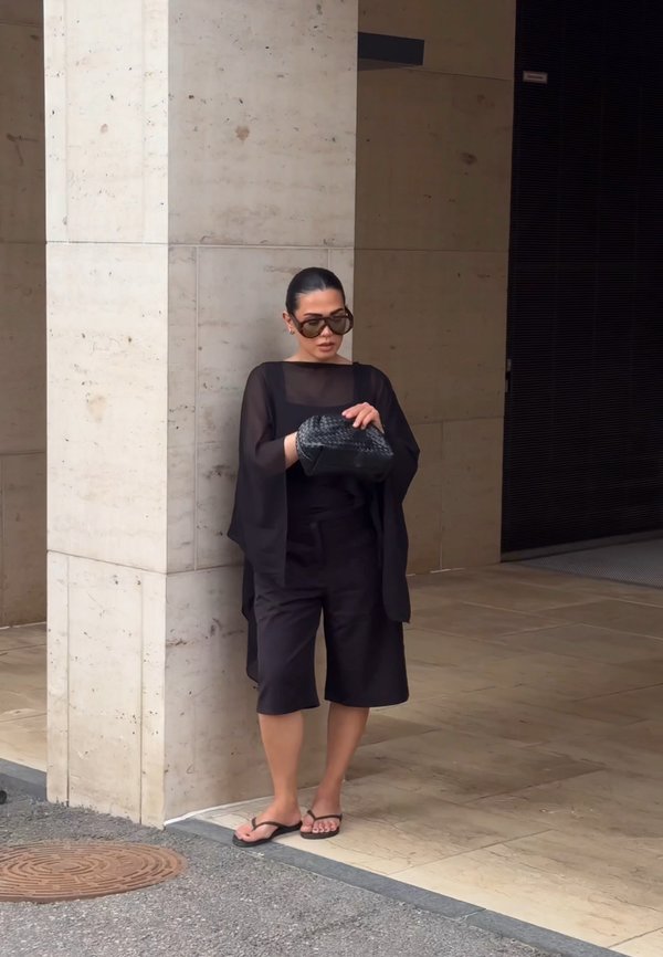 Frau in schwarzem Outfit, Sonnenbrille und Flip-Flops steht an einer Steinsäule und blickt in eine schwarze geflochtene Handtasche.