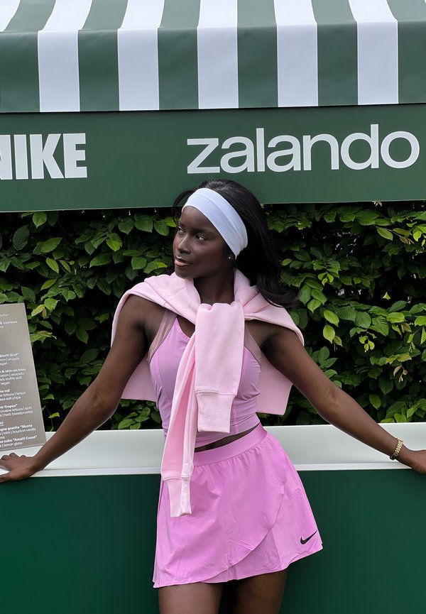 Femme sportive en tenue de sport rose et bandeau blanc appuyée contre une barrière verte sous un auvent rayé avec les logos Nike et Zalando.