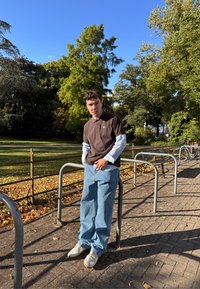 Mladik u slojevitoj košulji i polu-majici, naslonjen na držač bicikala pored park staze s palim lišćem ispod jasnog plavog neba.