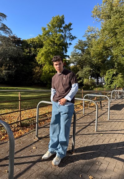 Joven con camisa de capas y polo, apoyado en un aparcamiento de bicicletas junto a un sendero del parque con hojas caídas bajo un cielo azul despejado.