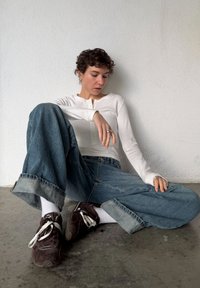 Personne avec des cheveux courts et bouclés portant un haut blanc à manches longues, un jean large, des chaussettes blanches et des baskets foncées, assise sur un sol en béton contre un mur blanc.