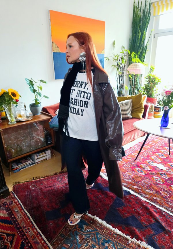 Red-haired woman wearing oversized coat, graphic t-shirt, and statement earrings walking in a colorful living room with plants and rugs.