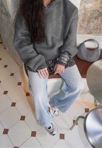 Personne assise sur un banc portant un sweat à capuche gris scintillant, un jean bleu clair, des baskets blanches, des bagues argentées, des bracelets, avec un sac à main gris à proximité.