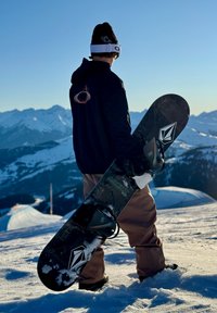 Snowboarder che tiene una tavola da snowboard nera e decorata con attacchi. Vestito con una giacca scura e pantaloni color sabbia, in piedi su una pista innevata con montagne sullo sfondo.