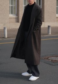 Casaco longo castanho escuro feito de lã, com aba larga e bolsos. Combinado com calças pretas e sapatilhas brancas numa superfície de rua.