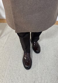 Personne portant des chaussures habillées sombres avec des glands, un pantalon noir et un manteau marron, debout sur un tapis texturé de couleur claire.