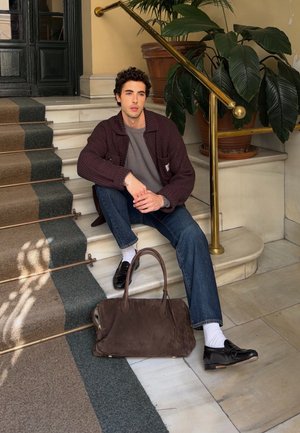 Joven sentado en escaleras de mármol junto a una barandilla de latón y una maceta grande, vistiendo un suéter oscuro, jeans y zapatos negros, con una bolsa marrón en el suelo.