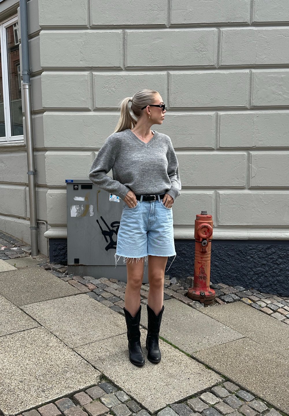 Siv jopica z V-izrezom, svetlo modri traper šortai s raztrganimi robovi, črne kavbojske škornje in sončna očala, stoji na teksturiranem tlaku.