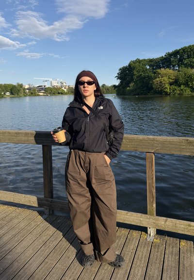 Joven mujer de cabello oscuro lleva chaqueta negra, pantalones anchos marrones, gafas de sol, cinta para la cabeza y sostiene una taza de café, de pie en un muelle de madera junto al lago.