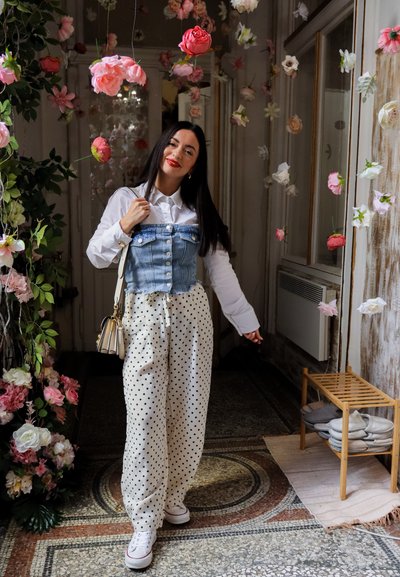 Mujer sonriente, de pie en interior bajo flores colgantes rosas y blancas, vistiendo camisa blanca, chaleco vaquero, pantalones de lunares y zapatillas blancas.