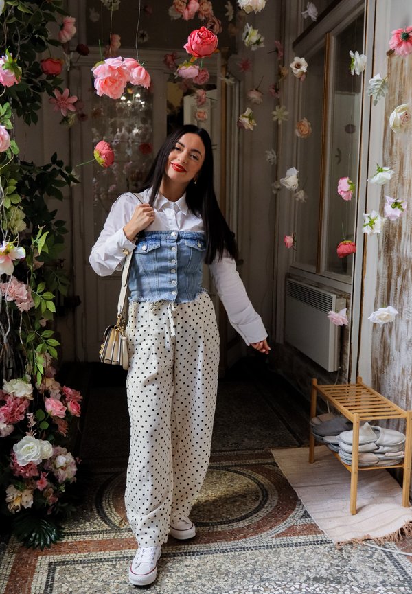 Femeie zâmbitoare, stând în interior sub flori roz și albe agățate, purtând cămașă albă, vestă din denim, pantaloni cu buline și adidași albi.