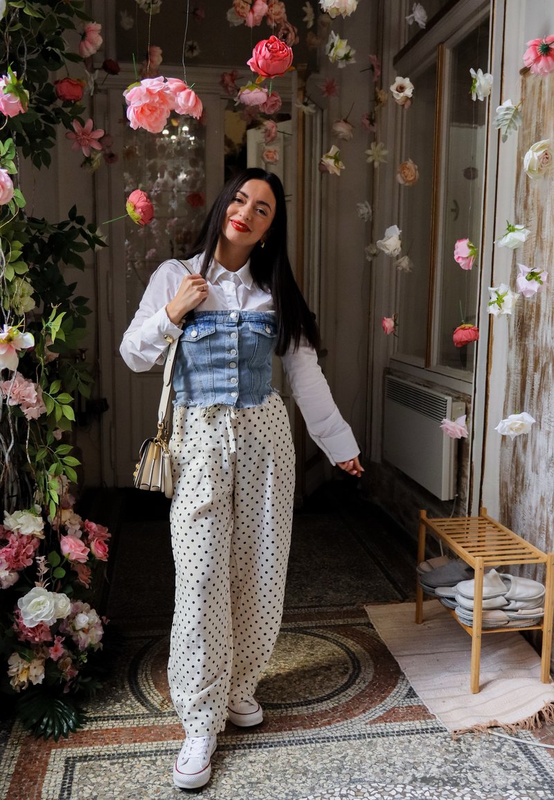 Γυναίκα που χαμογελά, στέκεται σε εσωτερικό χώρο κάτω από κρεμαστά ροζ και λευκά λουλούδια, φορά λευκό πουκάμισο, τζιν γιλέκο, πουά παντελόνι και λευκά αθλητικά παπούτσια.