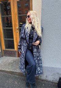 Μακρύ ναυτικό παλτό με λευκά γραφικά σχέδια, σε στρώσεις πάνω από denim σύνολο. Υφασμάτινη υφή και απλές μαύρες μπότες ολοκληρώνουν την εμφάνιση.