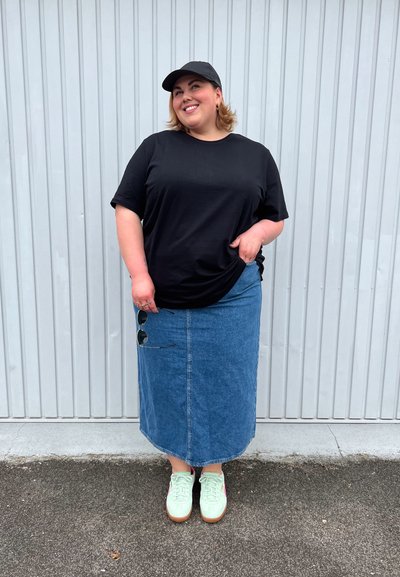 Persona sonriente que lleva una gorra negra y una camiseta, una falda de mezclilla azul, zapatillas deportivas verde claro, sosteniendo gafas de sol, de pie frente a una pared de cartón ondulado gris.